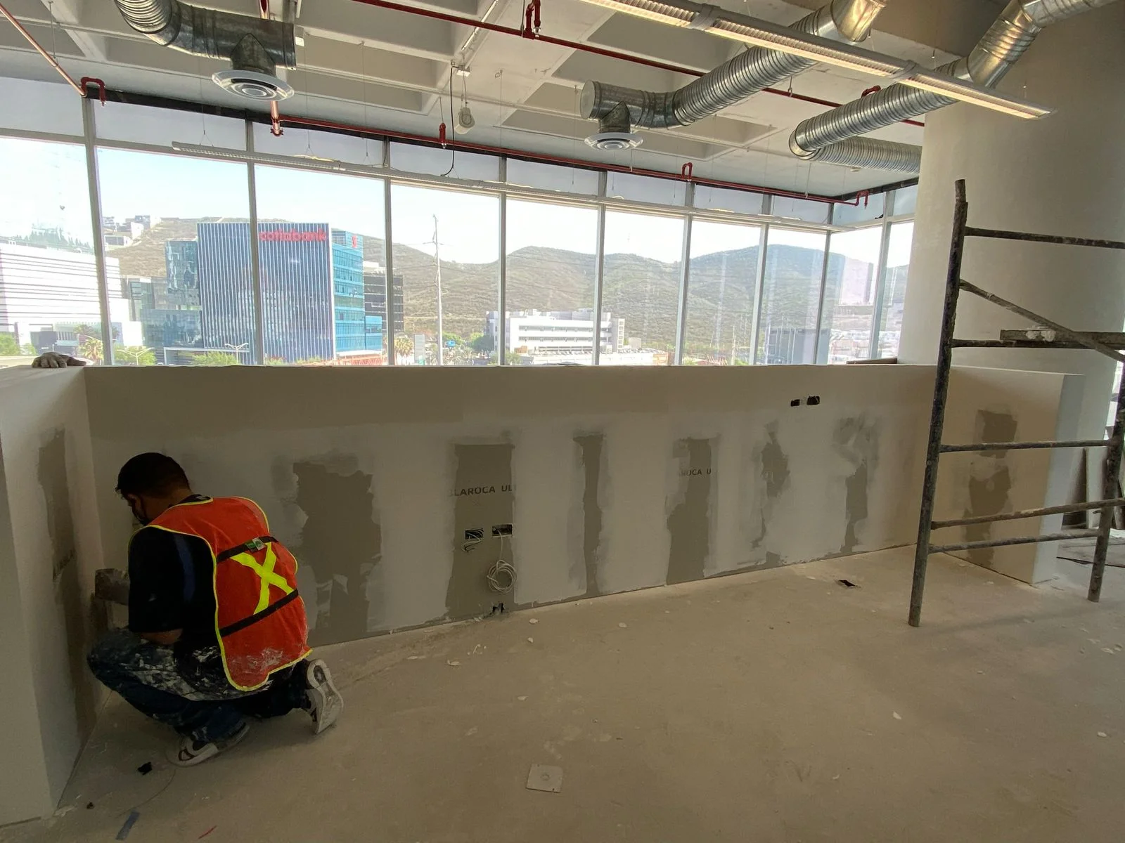 Trabajador resanando muro de tablaroca con vista panorámica al skyline corporativo — Nórdika Business Center Tempus